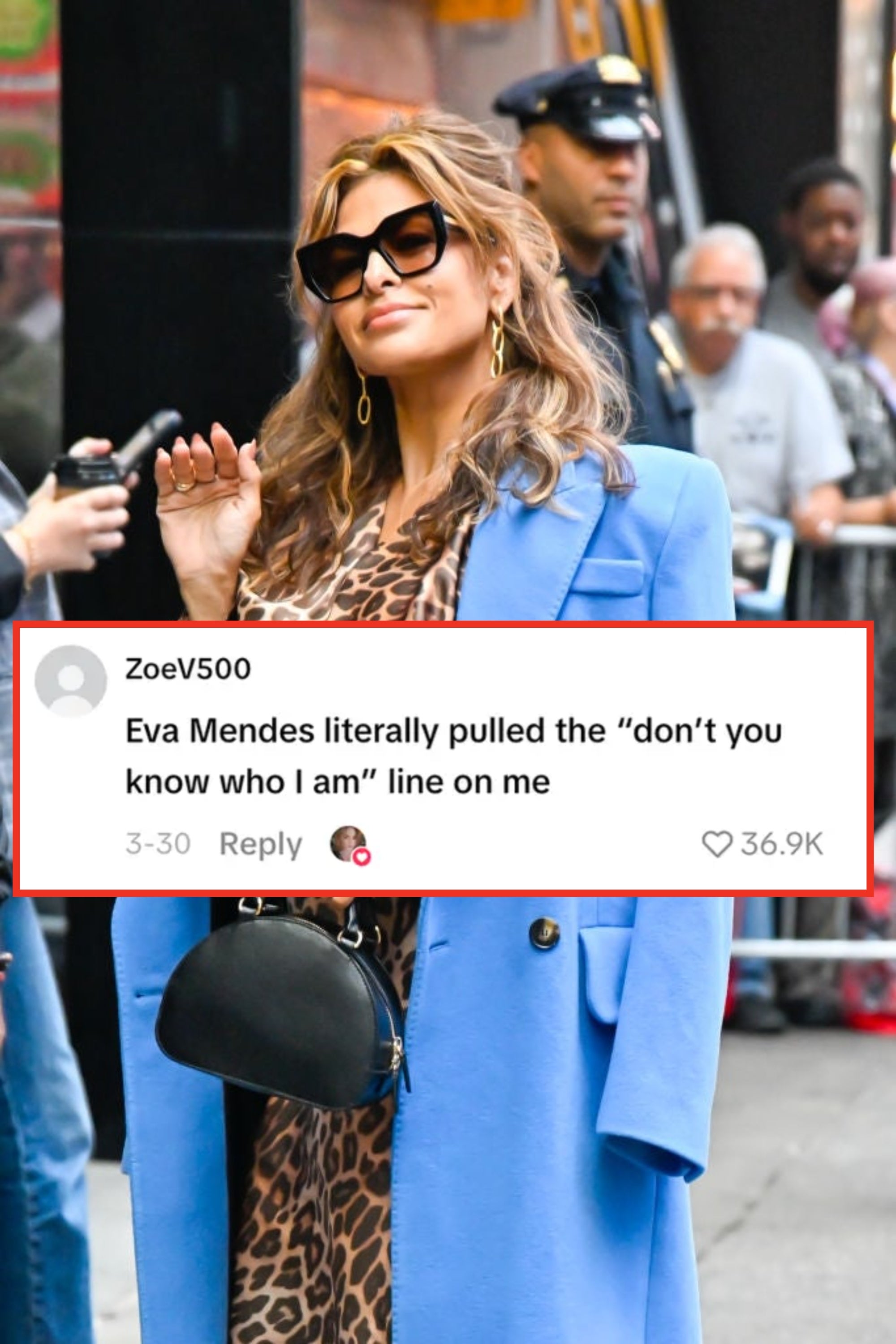 Celebrity in a leopard print dress with a blue coat, holding a black handbag and wearing sunglasses, waves while outdoors