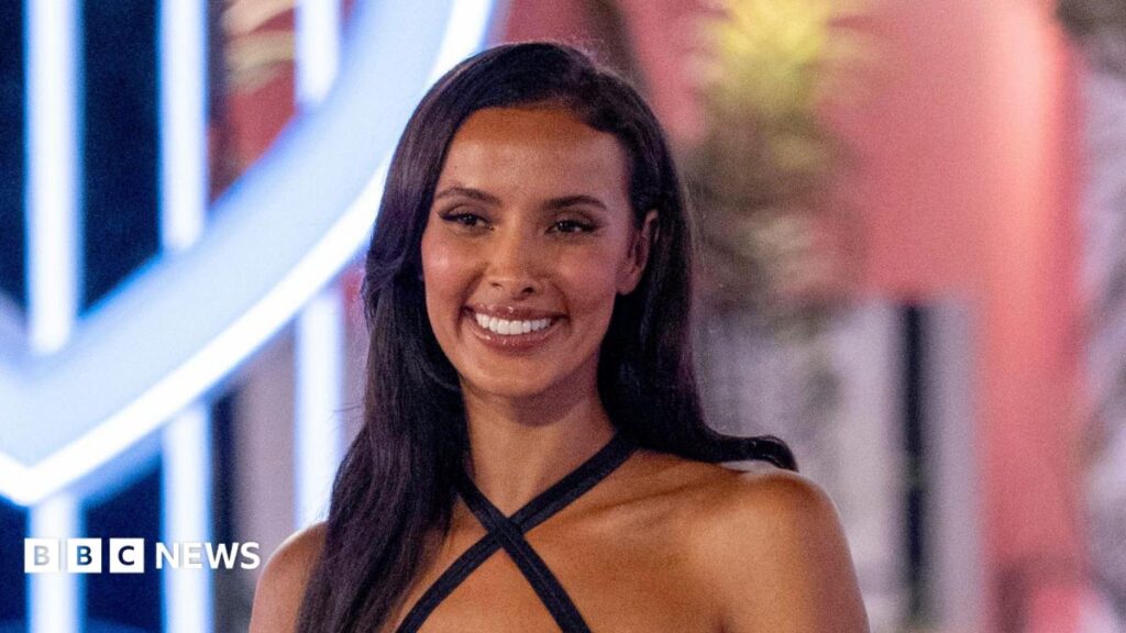 A woman with short brown hair looks directly into the camera with a slight smiling expression. She is wearing a pink top and a silver necklace with a heart charm.