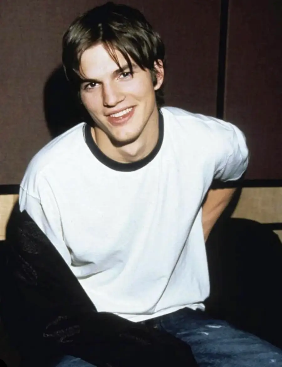 A young man with brown hair smiles while sitting, wearing a white T-shirt with black trim and blue jeans, in front of a dark-colored background.