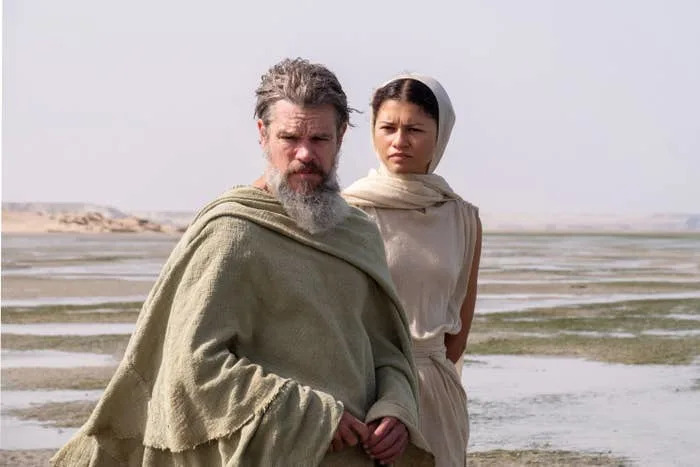 Two actors stand in a desolate landscape, dressed in draped, rustic attire, evoking a historical or fantasy setting