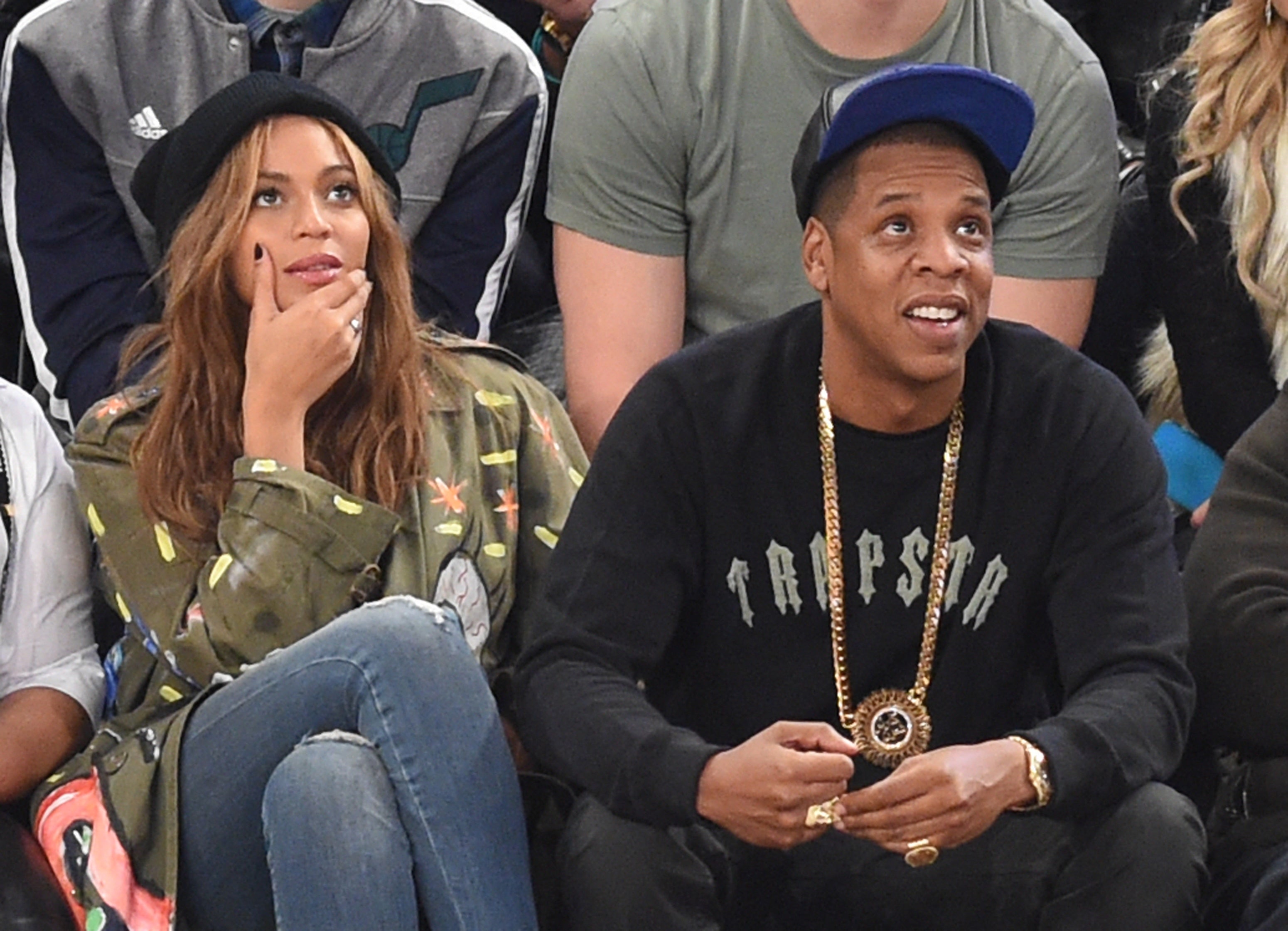 Beyoncé and Jay-Z sit beside each other at a sports game. Beyoncé wears a camo jacket and blue jeans while Jay-Z wears a black hoodie and gold chain