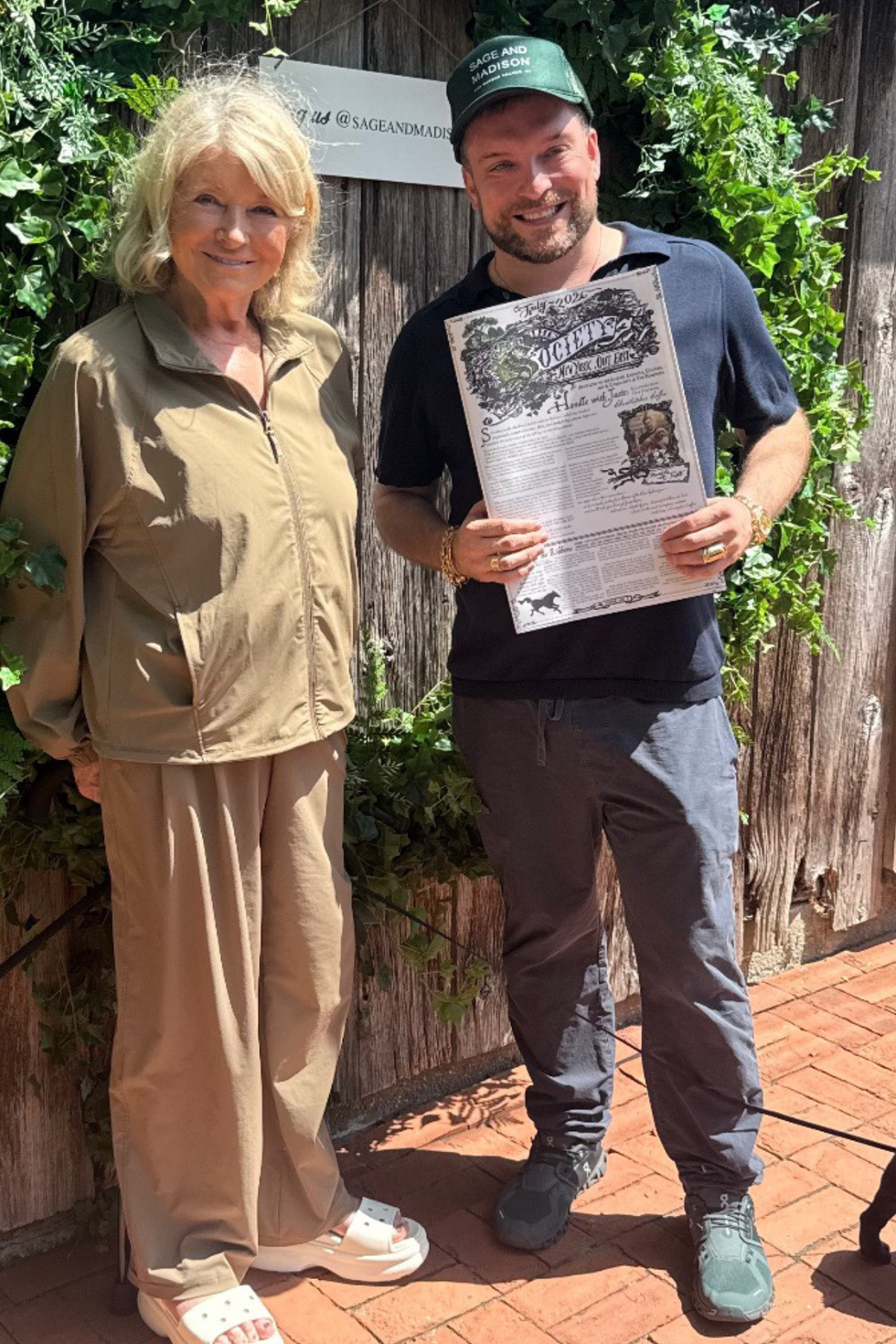 Two individuals standing together outdoors, one holding a printed document, surrounded by greenery.