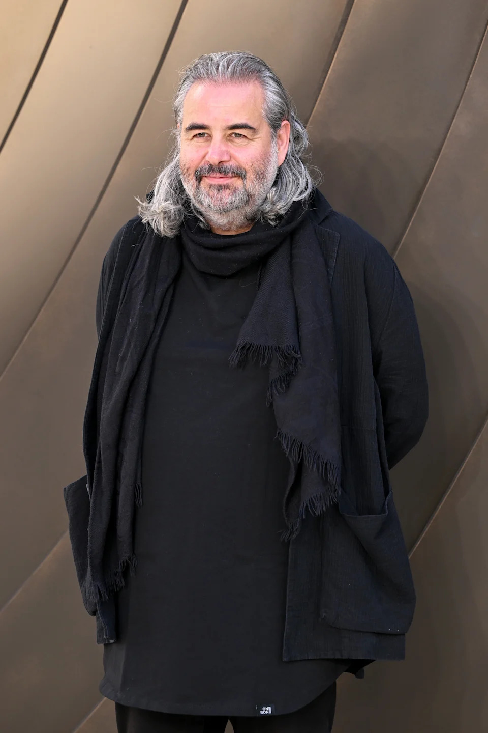 Person with long gray hair and beard, wearing a layered black outfit, stands against a patterned backdrop