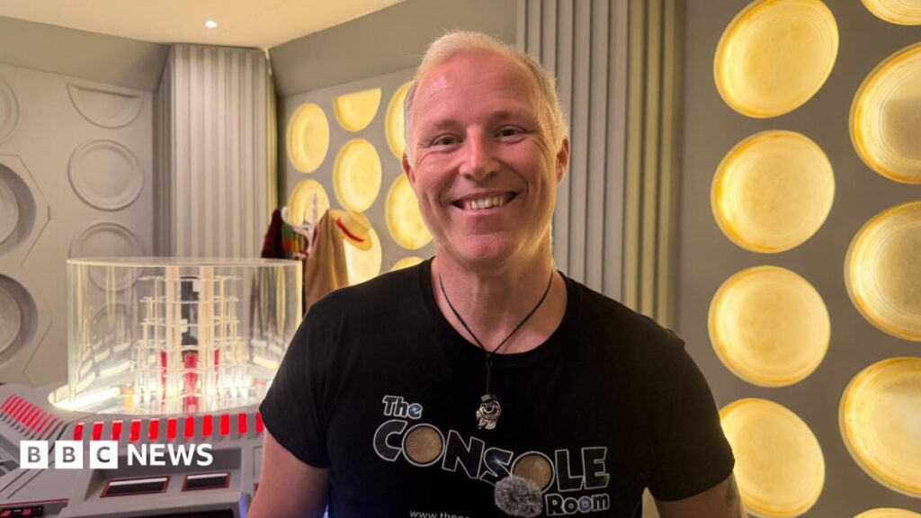 Doctor Who fan builds 1983 Tardis console room in his back garden A man with short blond hair is smiling for a head and shoulders photo and is wearing a black top that says The Console Room on it. He is also wearing a circular necklace on a black thin rope. Behind him are grey walls with yellow circles on it, a hat stand with a hat and brown coat and a scarf on the other side. Next to him is a clear cube on a console with several buttons.
