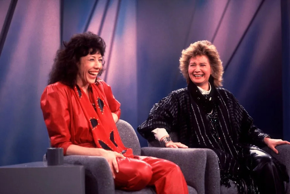 Two people are sitting and laughing on a talk show set. One wears a bold, loose-fitting outfit, and the other is in a textured, layered ensemble