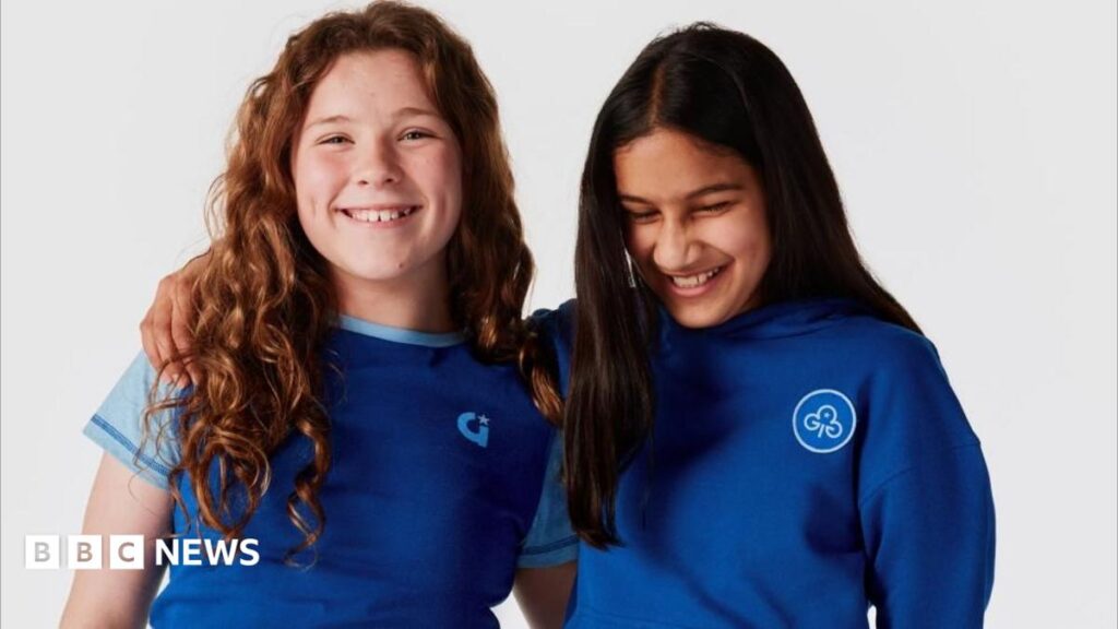 Two girls stand next to one another with their arms around the other's shoulder. The one on the left has curly ginger hair while the one on the right has dark, straight hair. They are both smiling. The girl on the left is wearing a short sleeved dark blue top with light blue cap sleeves and a small Girlguiding logo on the front. The other girl is wearing a royal blue hoodie with a circular Girlguiding logo patch on the front. The background is plain white.