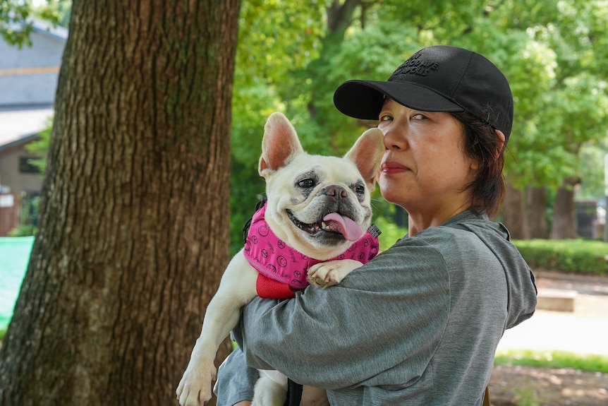 A woman in a grey hoodie and black cap holds a white pug dog in a pink jacket.