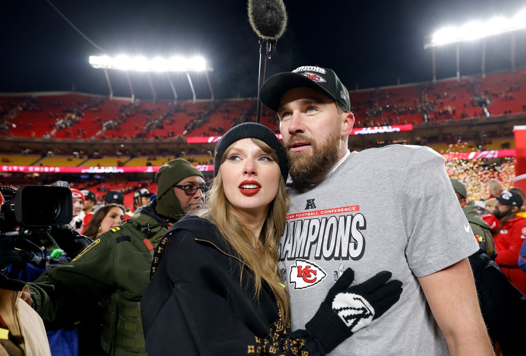 Taylor Swift wearing a black beanie and a dark jacket with gold accents hugging Travis Kelce, who is wearing a gray AFC Champions t-shirt and a Kansas City Chiefs cap.