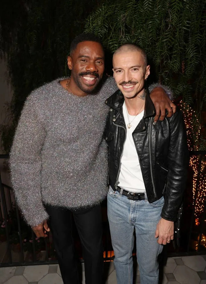 Two people smiling, one in a textured sweater, the other in a leather jacket with jeans, pose together at an event