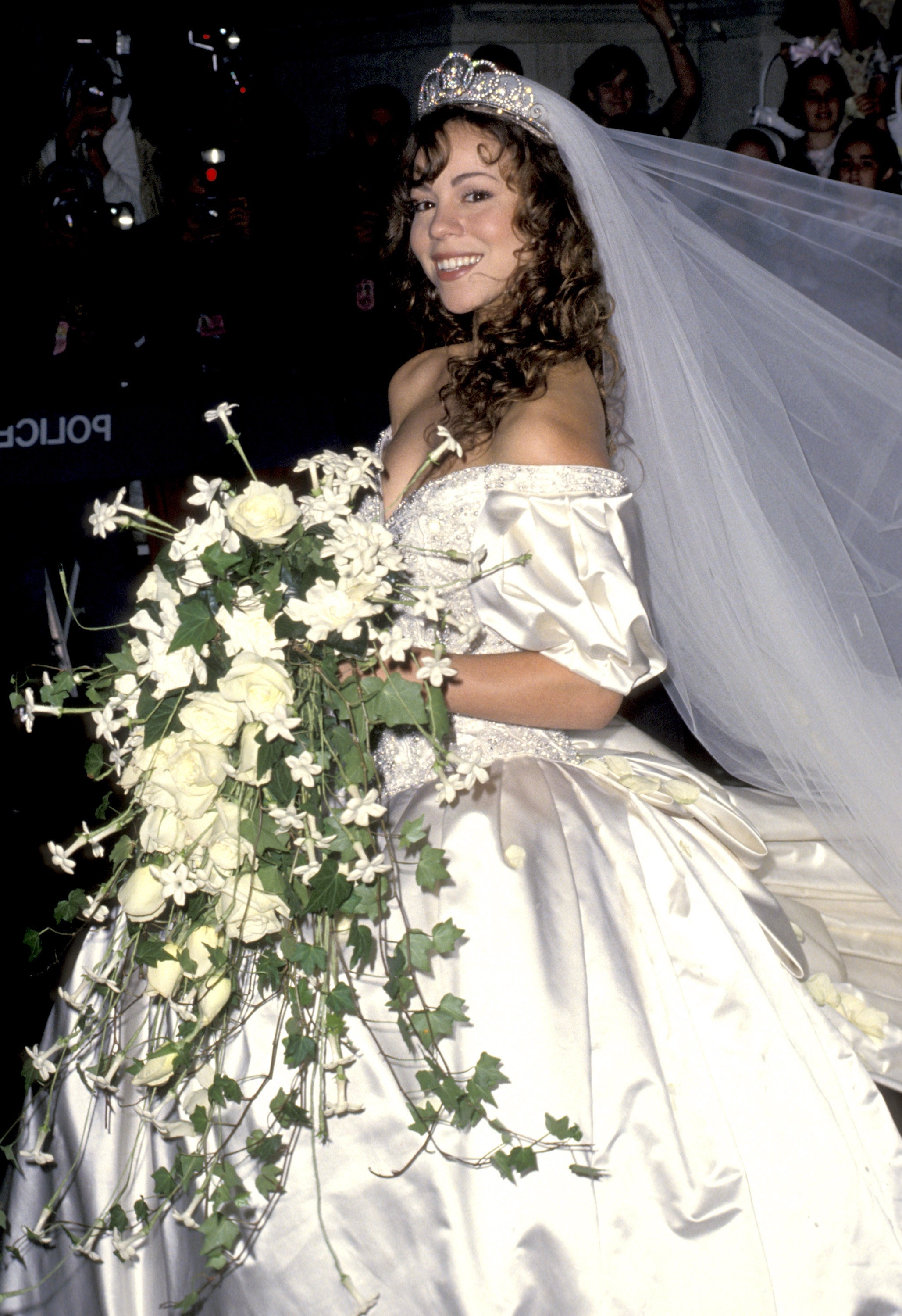 A celebrity in an elaborate wedding gown with a long veil and tiara, smiling while holding a large bouquet of white flowers