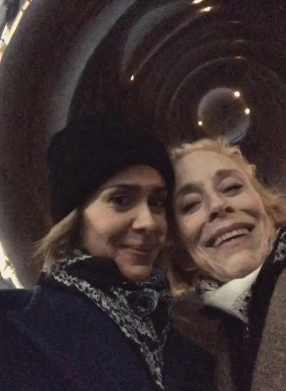 Two individuals are smiling for a selfie, bundled up in winter clothing, one wearing a beanie, against a dimly lit, tunnel-like background
