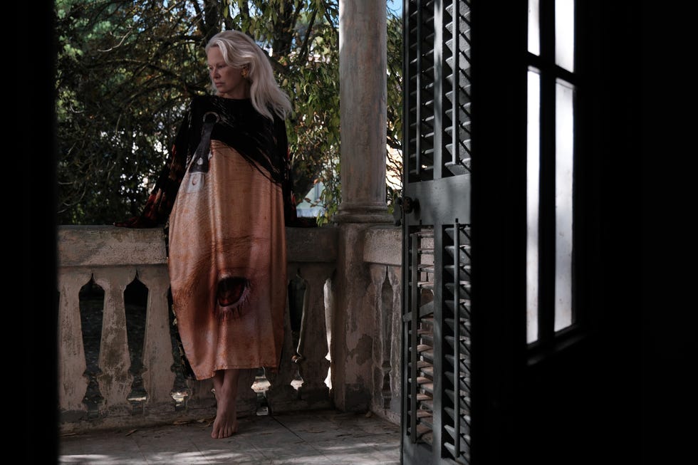 A woman stands on a balcony wearing a long artistic dress with a striking design, surrounded by greenery and natural light.