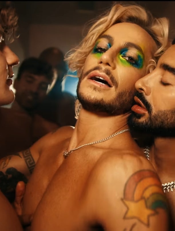 Three individuals are closely gathered; one with colorful eye makeup, another wearing a chain necklace. All are shirtless, in an intimate setting