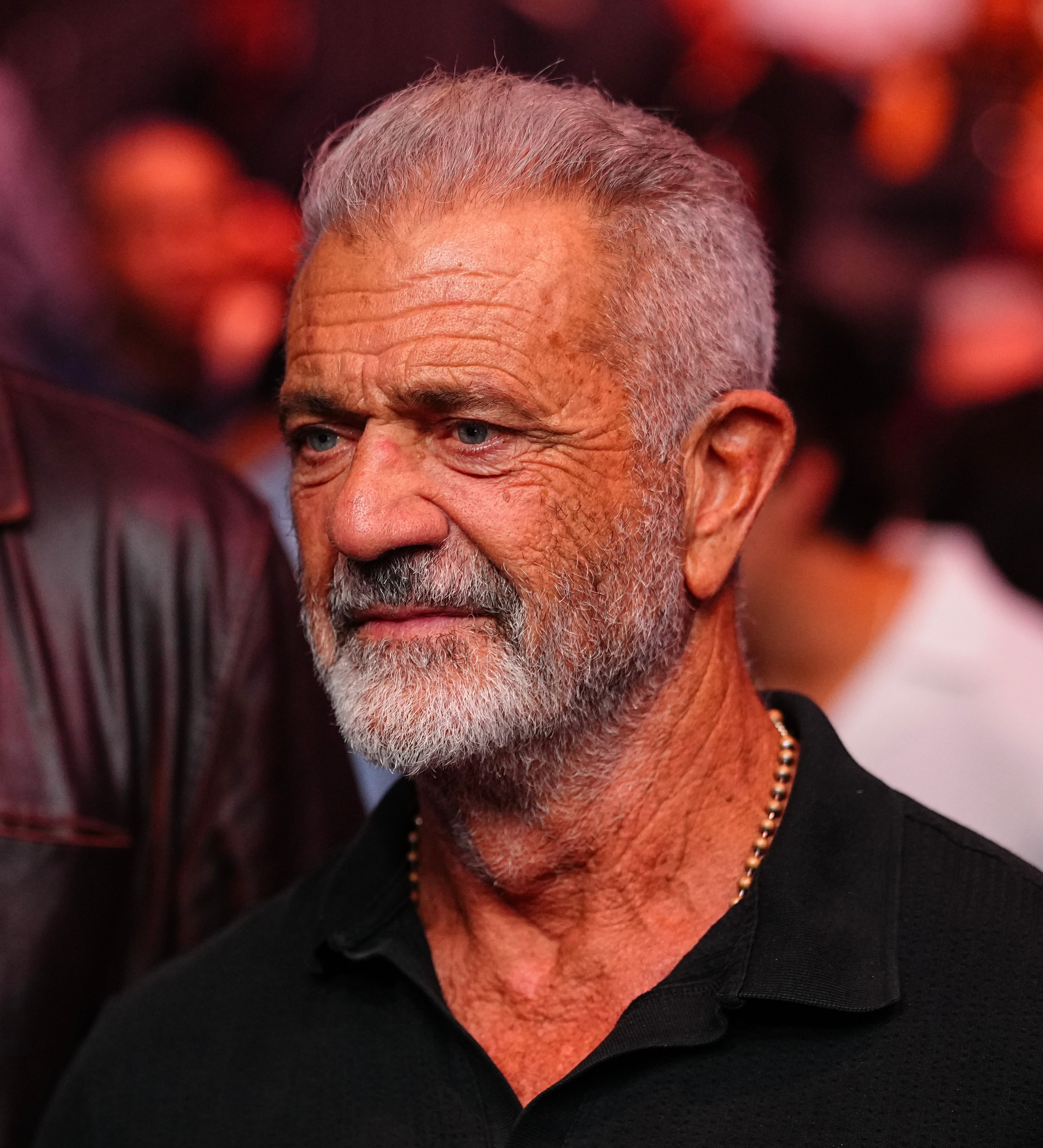 Mel Gibson in a casual polo shirt stands at an event, appearing engaged with the surroundings