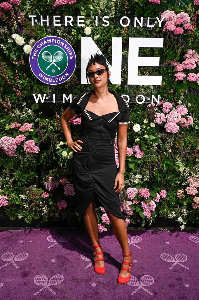 Joy Crookes in a black dress and red heels stands in front of a floral Wimbledon backdrop.