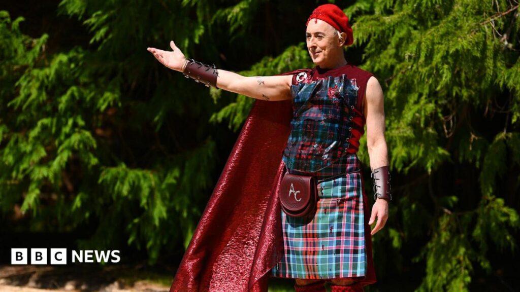 Traitors US host Alan Cumming donates outfit to V&A Dundee Alan Cumming, a man, is stood holding his arm out with trees behind him. He has a red beret on his head, a leather wrist band, a red cape, a tartan vest, tartan kilt, red knee high socks and a sporran which have the initials AC embroidered onto them. He is smirking slightly and looks as if he is welcoming people to something.