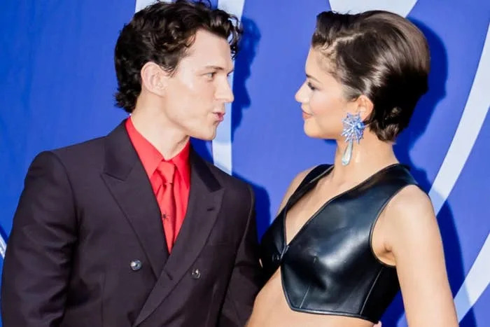 Two people on a Spider-Man-themed red carpet. One wears a dark suit with a red shirt; the other wears a black leather crop top and skirt