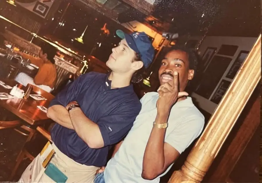 Two men pose inside a restaurant; one stands with arms crossed looking sideways, wearing a blue shirt and cap. The other smiles, pointing at the camera, wearing a dark shirt and watch. Tables and diners are in the background.