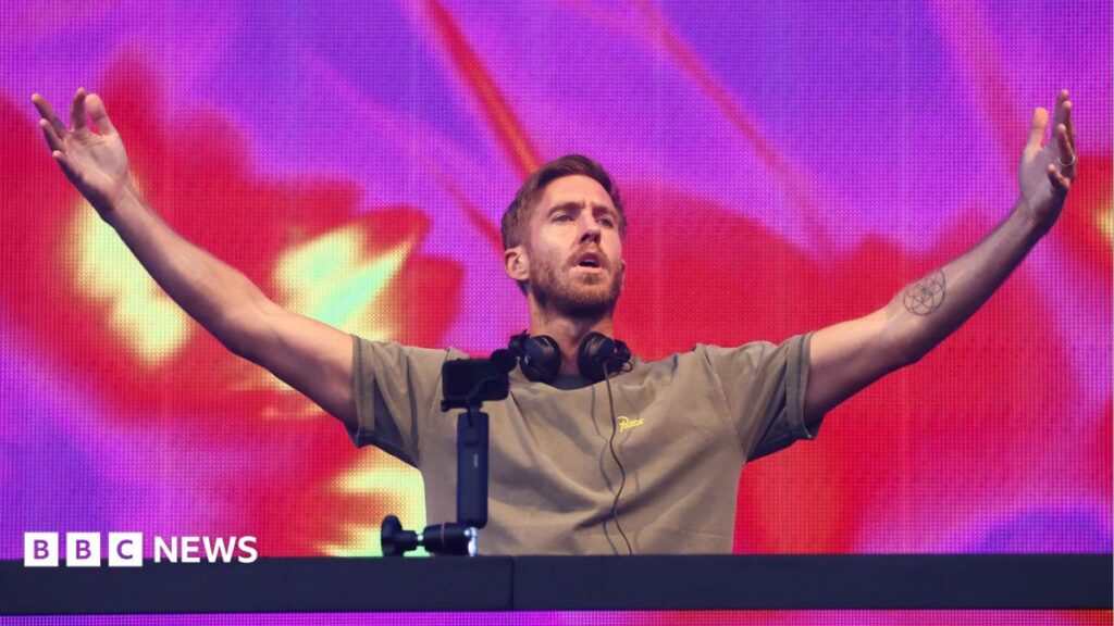 Calvin Harris homecoming show to be broadcast live on BBC Calvin Harris wears a plain grey T-shirt and stands at the DJ booth on stage. He has headphones around his neck and holds his hands up in the air. Behind him you can see a screen with purple, red and yellow shapes