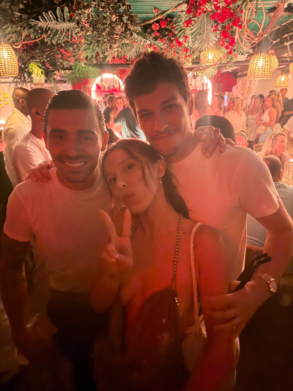 Three friends smiling and posing together in a lively, decorated venue with dim lighting and plants in the background.