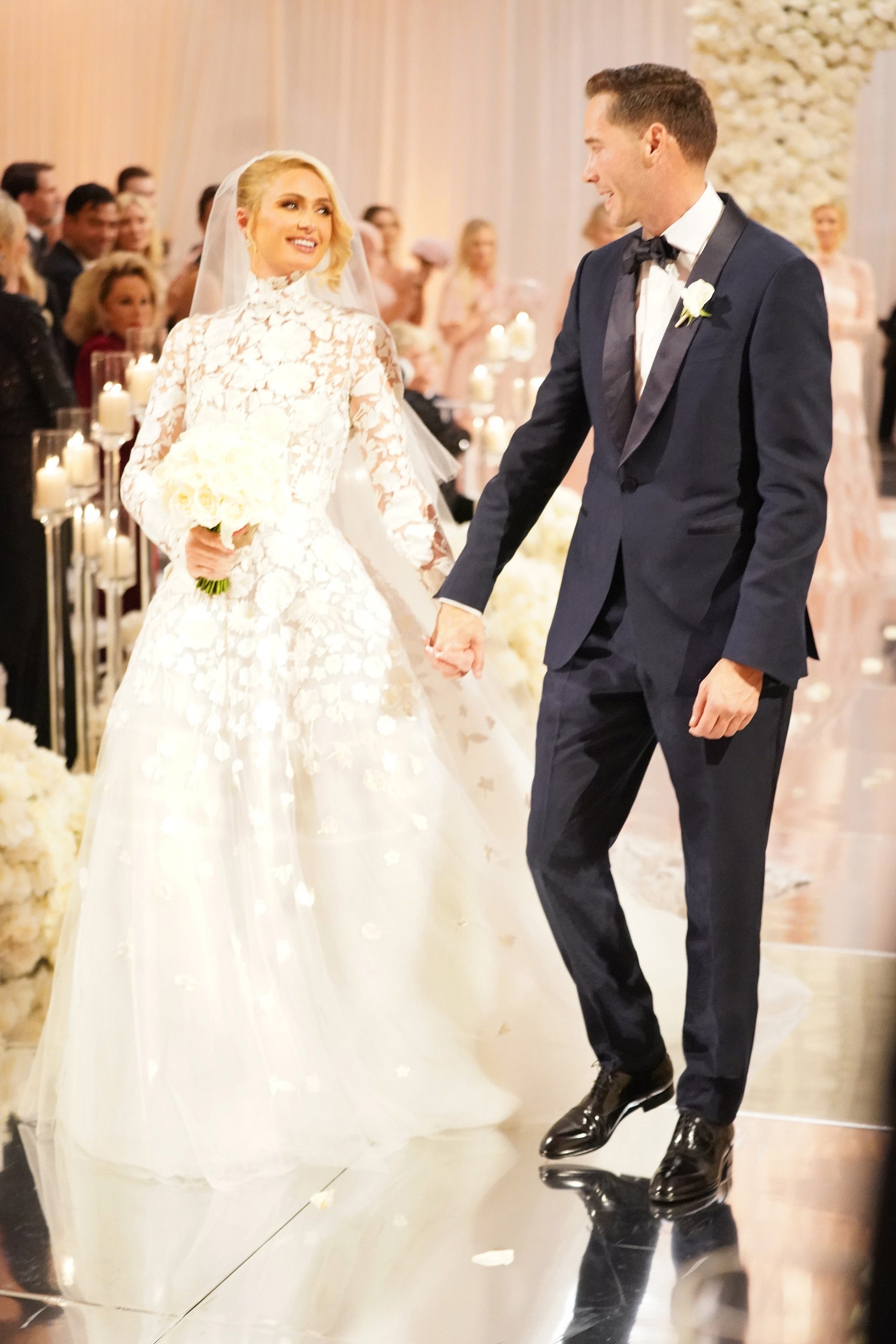 A couple walks an aisle; the woman wears a floral lace gown and veil, holding a bouquet. The man is in a tuxedo with a bow tie
