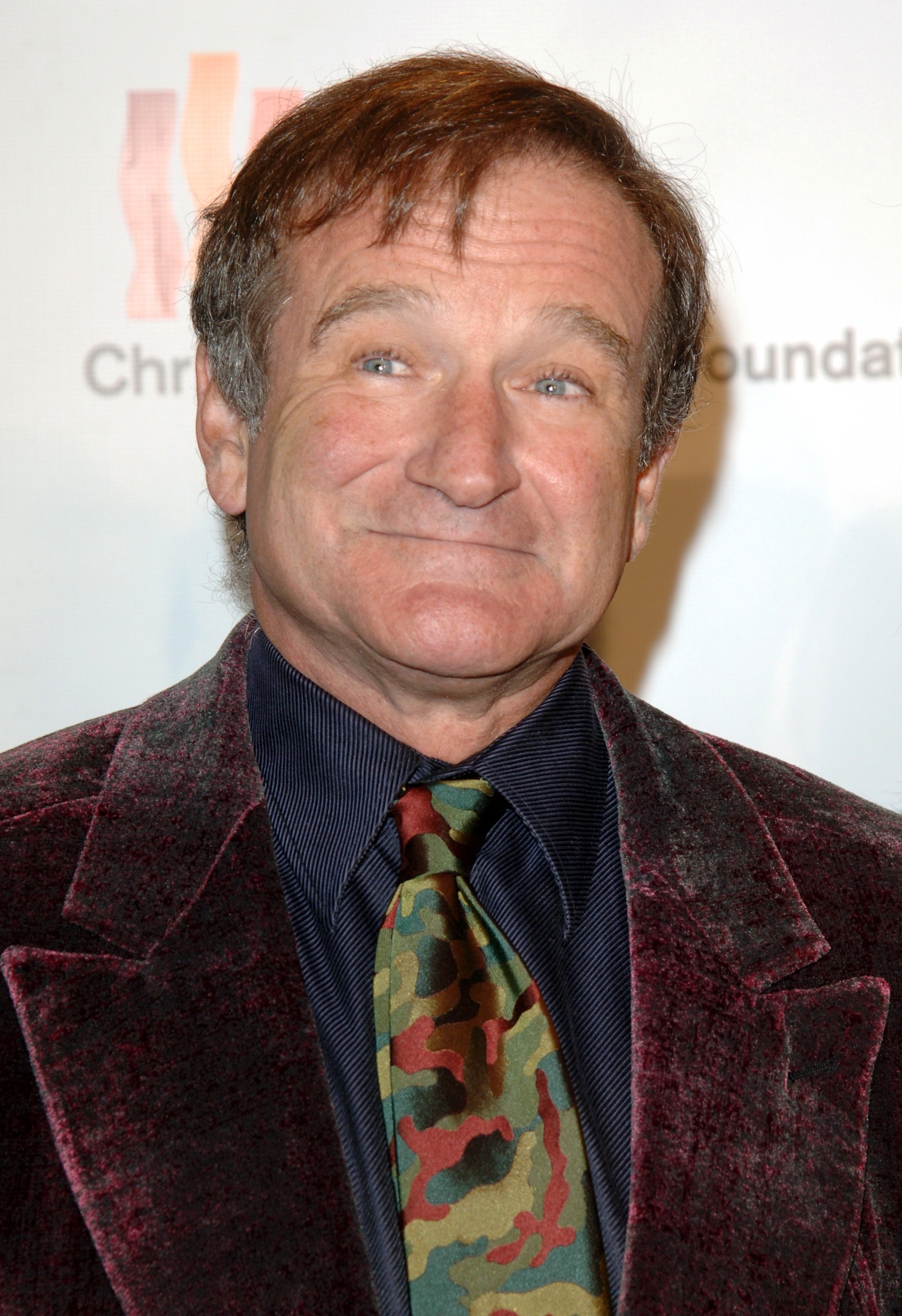 Robin Williams wearing a velvet jacket, a striped shirt, and a camouflage tie, smiling at an event