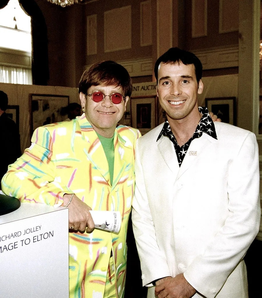 Two people pose together. The person on the left wears a vibrant suit with a bright shirt, and the person on the right wears a classic white suit