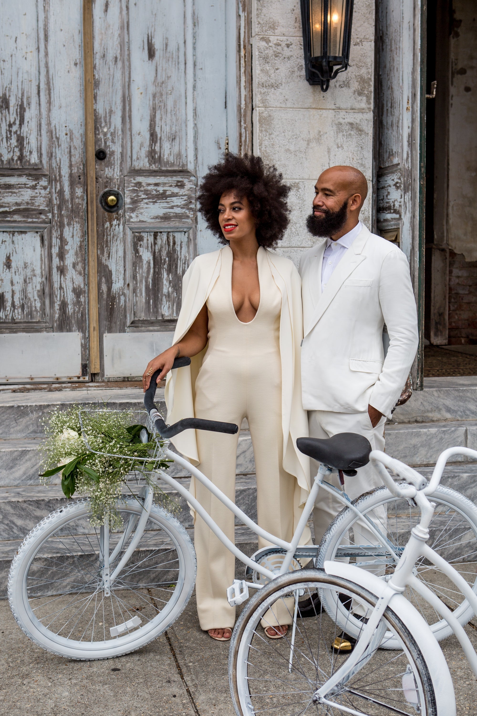 Two people stand with white bikes; one wears a cream jumpsuit with a cape, the other a white suit. They're outside an old wooden door