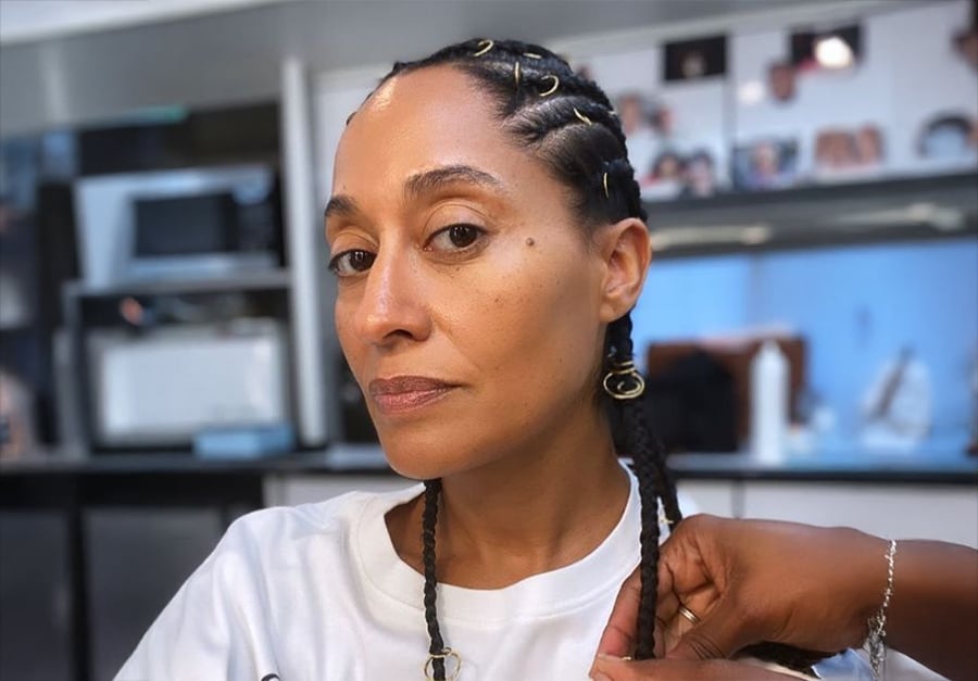 A woman with cornrow braids, adorned with gold hair cuffs, sits in a salon while someone styles her hair. She wears a white shirt and looks confidently at the camera. The background shows salon equipment and photos.