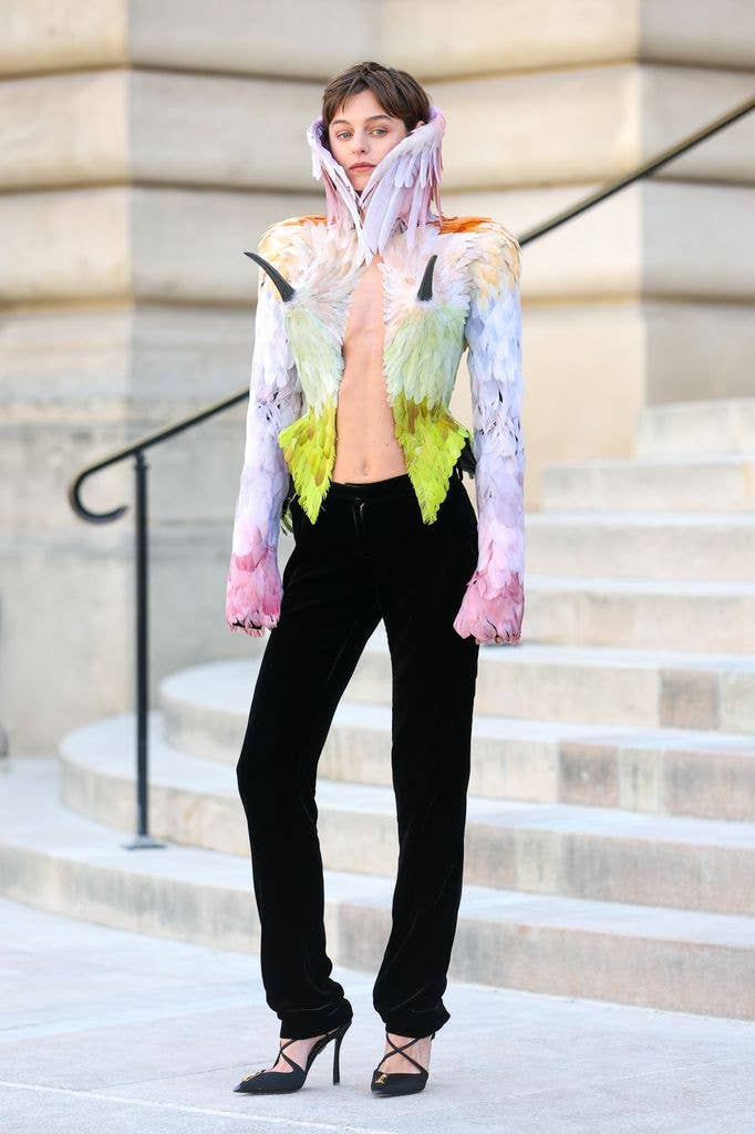 Emma Corrin  wears a multi coloured feathered jacket and black trousers
