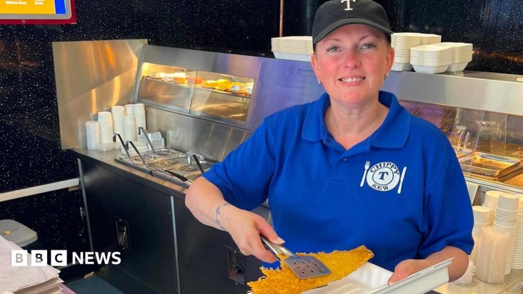 Fish and chip shop staff say working in heatwave is ‘horrendous’ Tracy Stock with hair tied back wearing black baseball cap and a blue t-shirt stands with a white Chippy-T logo stands in front of the frying range in a fish and chip shop holding a battered fish over a take out container with stainless steel tongs. She is smiling.