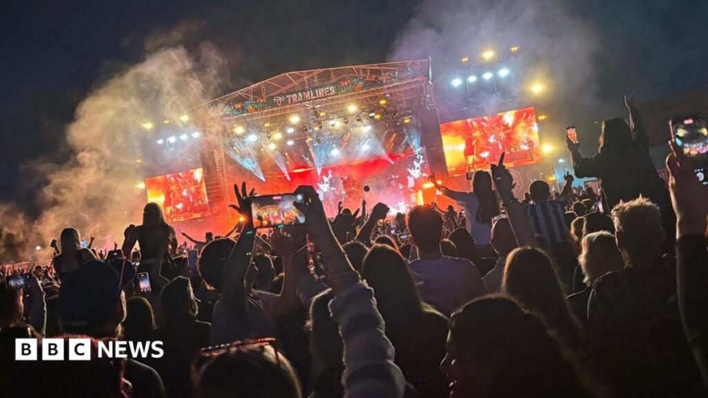 A large crowd scene at night with hundreds of music fans facing a large stage lit by pink lighting