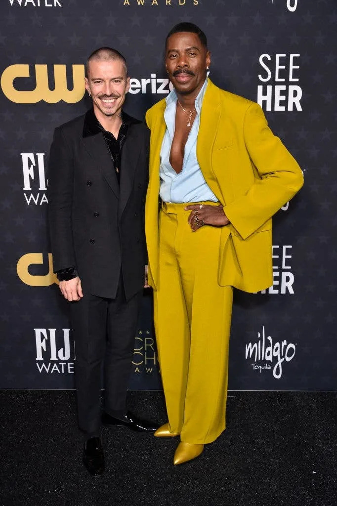 Two individuals on a red carpet; one in a black suit, the other in a vibrant yellow suit with an open shirt, at an award event