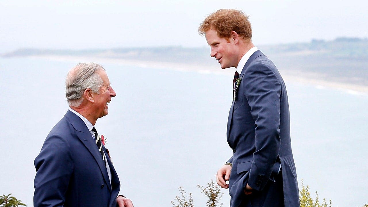 King Charles and a younger Prince Harry wearing matching dark blue suits in conversation and smiling.