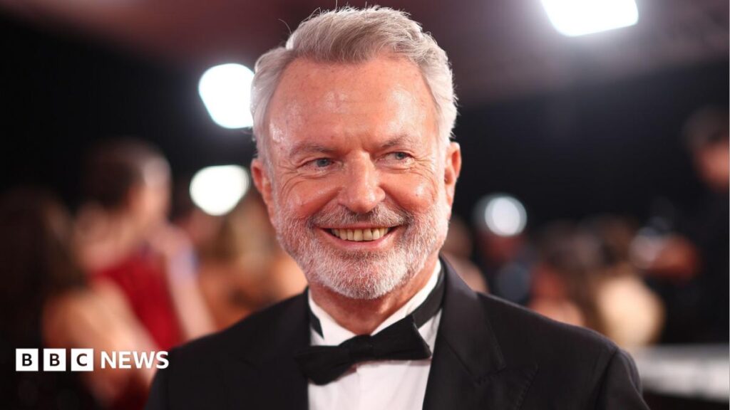 Sam Neill: New Zealand actor Sam Neill died from pneumonia, agent confirms Graham Norton has white hair, a moustache and beard. He is wearing a dark-colour T-shirt and sits behind a microphone with a green muffler.