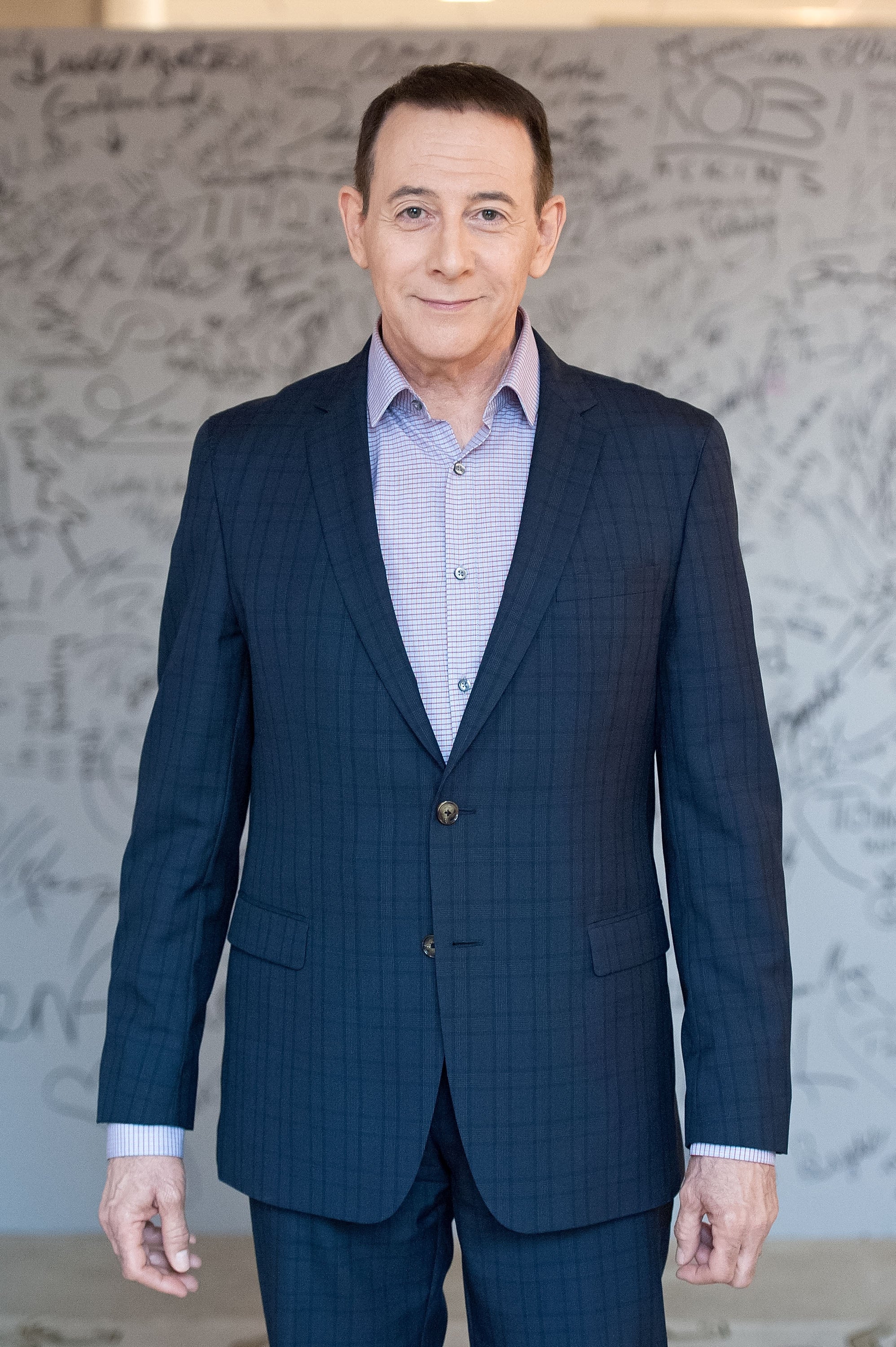 Paul Reubens in a checkered suit and open-collared shirt poses in front of a graffiti-covered wall, smiling gently