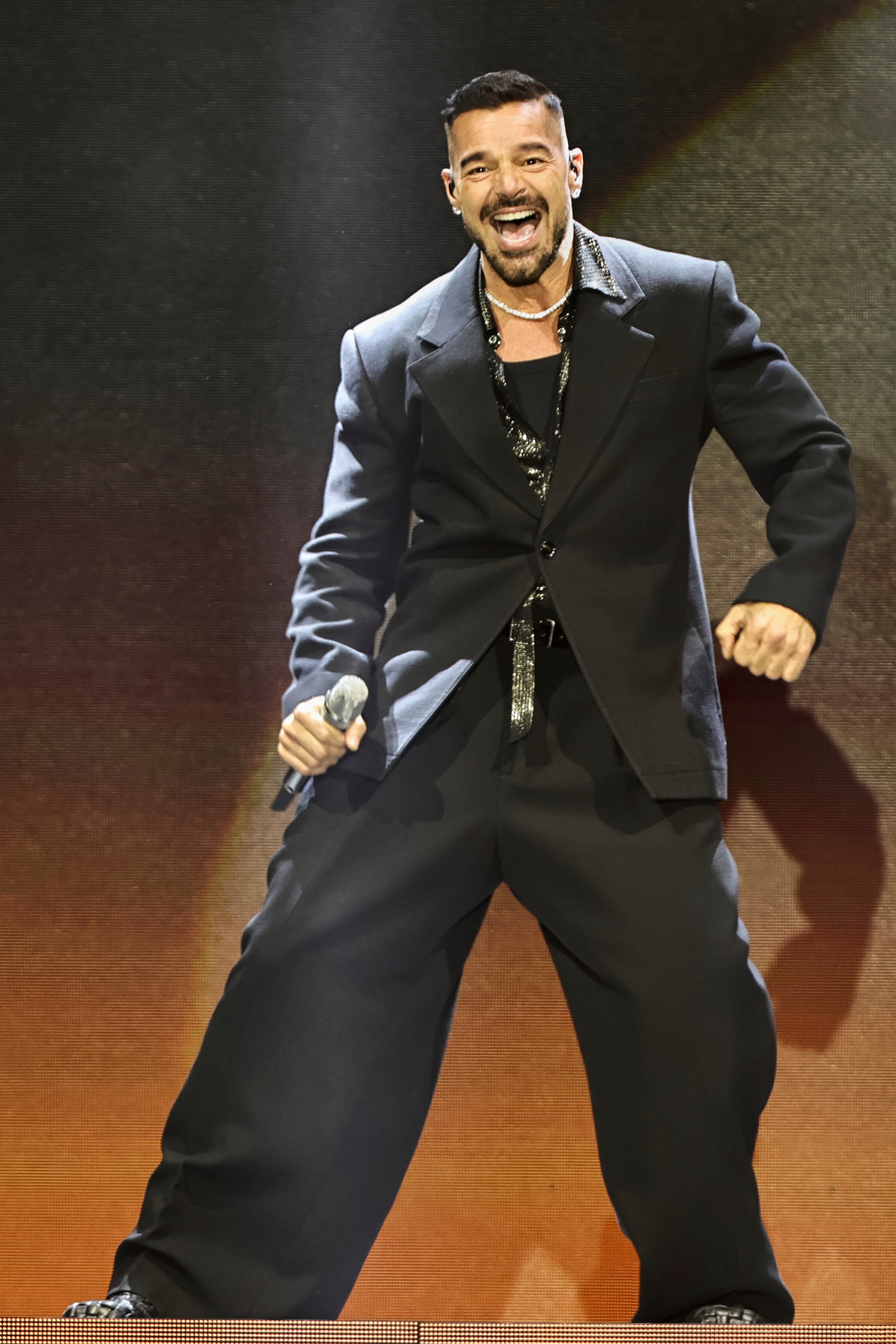 Singer energetically performing on stage, wearing a loose-fitting black suit with a shimmering shirt and a microphone in hand
