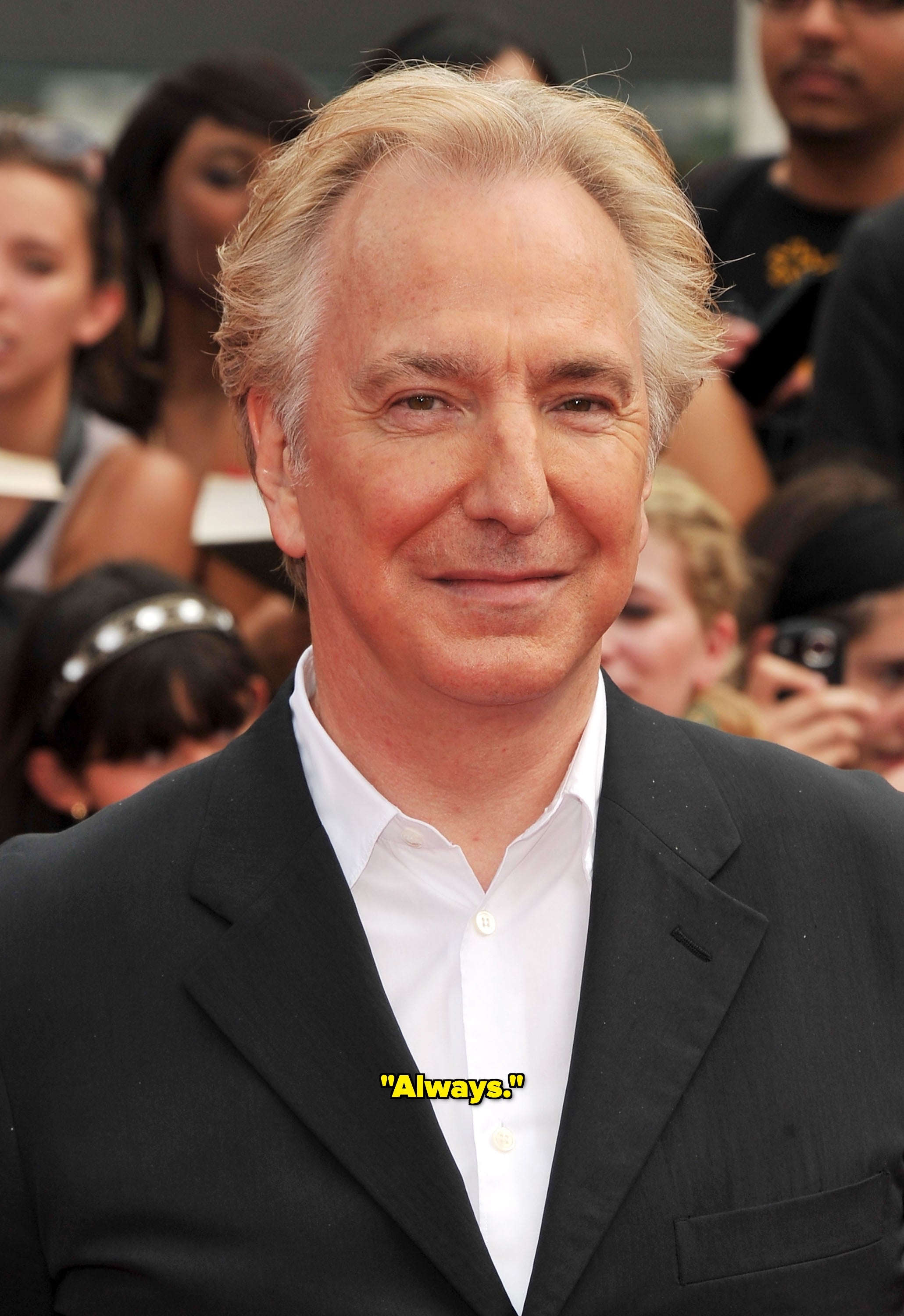 A person in a black suit and white shirt smiles at an outdoor event with people in the background