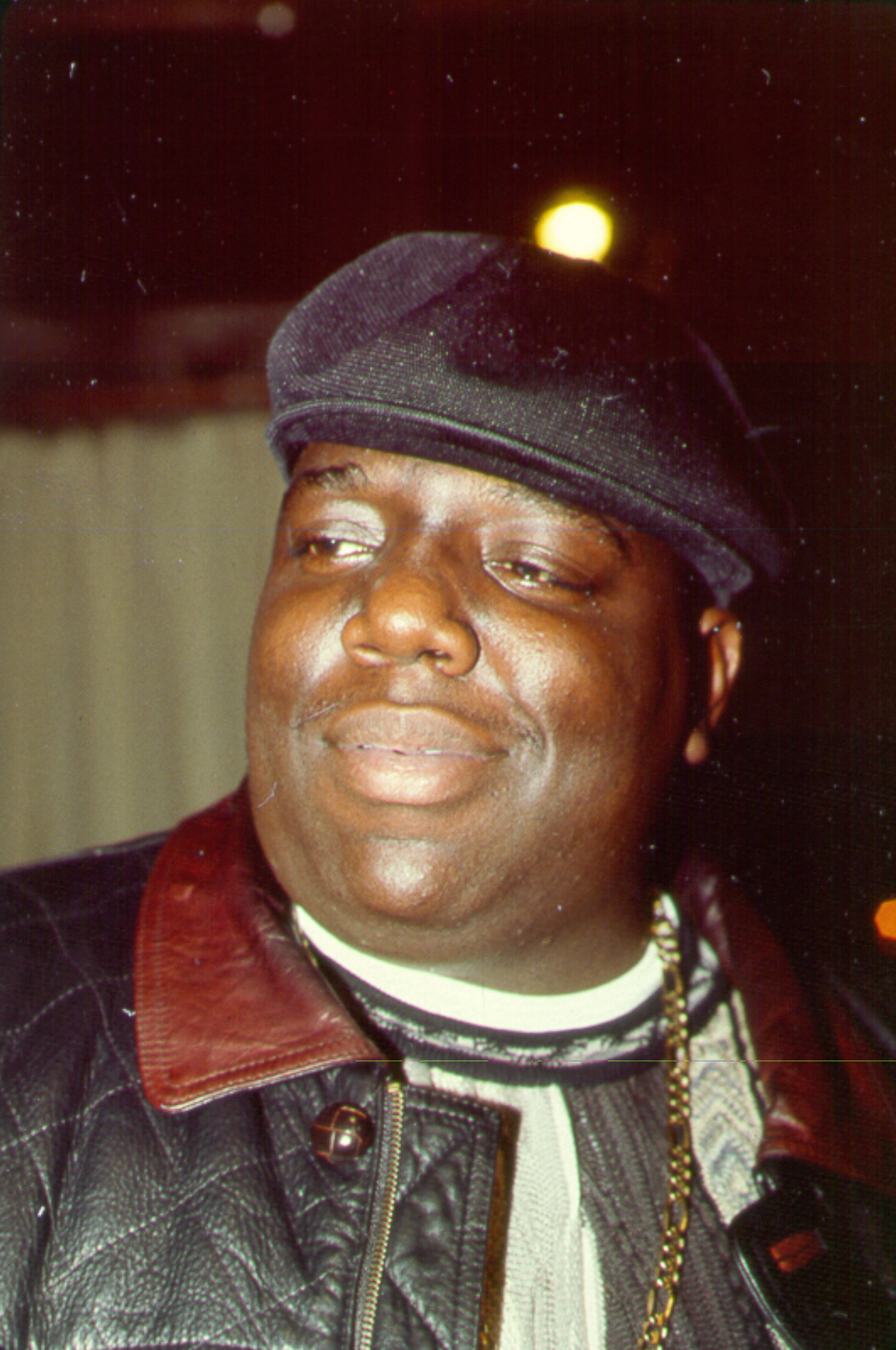 Biggie Smalls wears a flat cap and leather jacket with a thoughtful expression