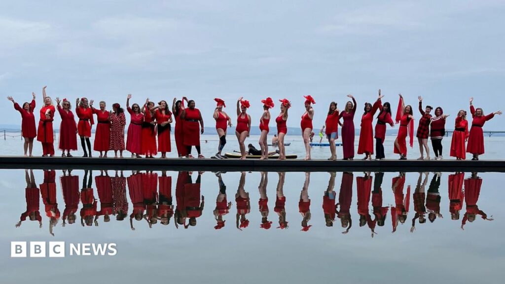 A long line of women wearing red dresses photographed standing next to a lake, with their reflection immediately below in the water.