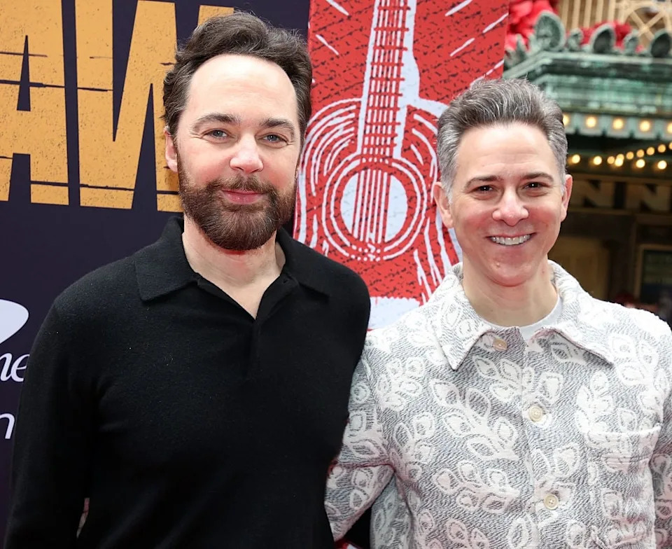 Two people pose together on a red carpet. One wears a black sweater, the other a patterned jacket. The backdrop has stylized graphics