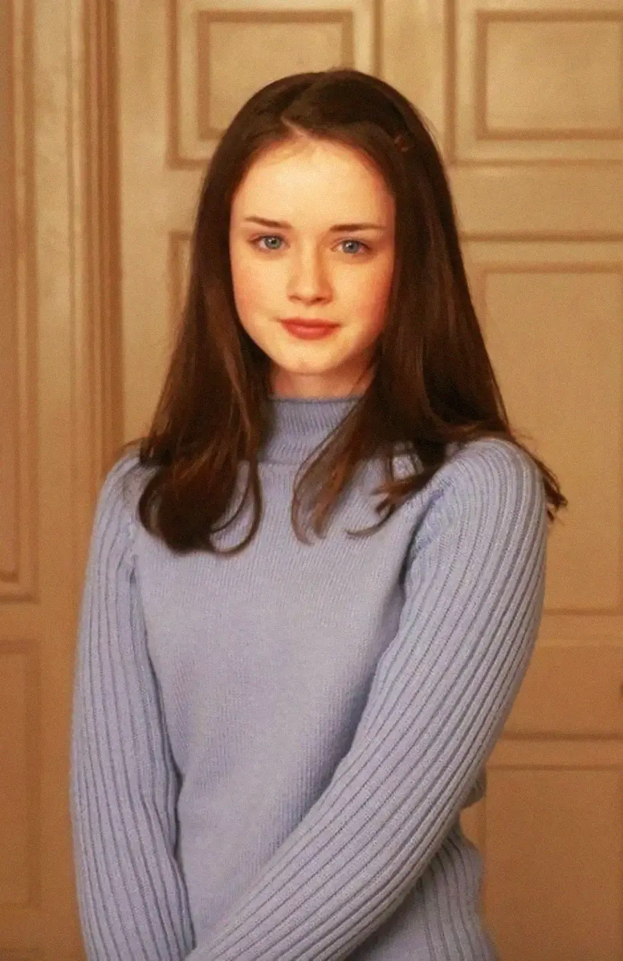 A young woman with long brown hair, wearing a light blue ribbed sweater, stands indoors in front of beige paneled doors, looking calmly at the camera.