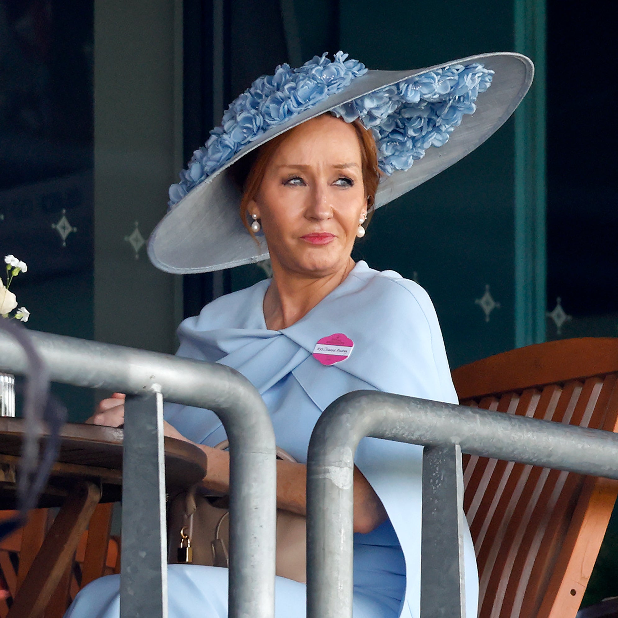 J.K. Rowling in elegant attire with a large, floral hat sits outdoors at a table, gazing thoughtfully