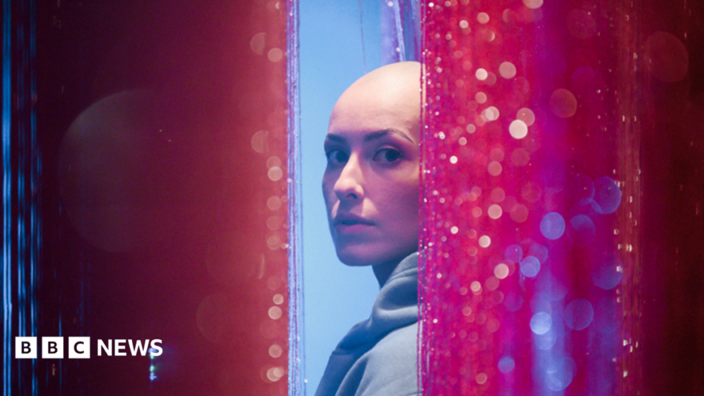 ‘I never dreamed I’d go on stage completely bald’ Emily Glaze is turning to look at the camera side-on from between pink glittery curtains. She has a bald head and is wearing a light blue hoodie.