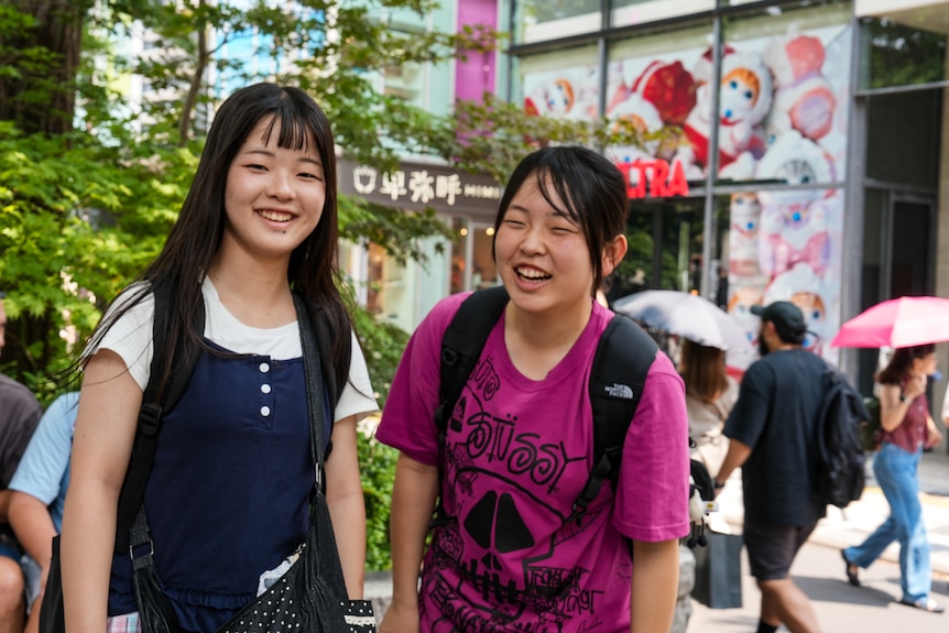 Two teenage girls smiling and laughing, while pedestrians carrying sun parasols stroll past on the street behind them.