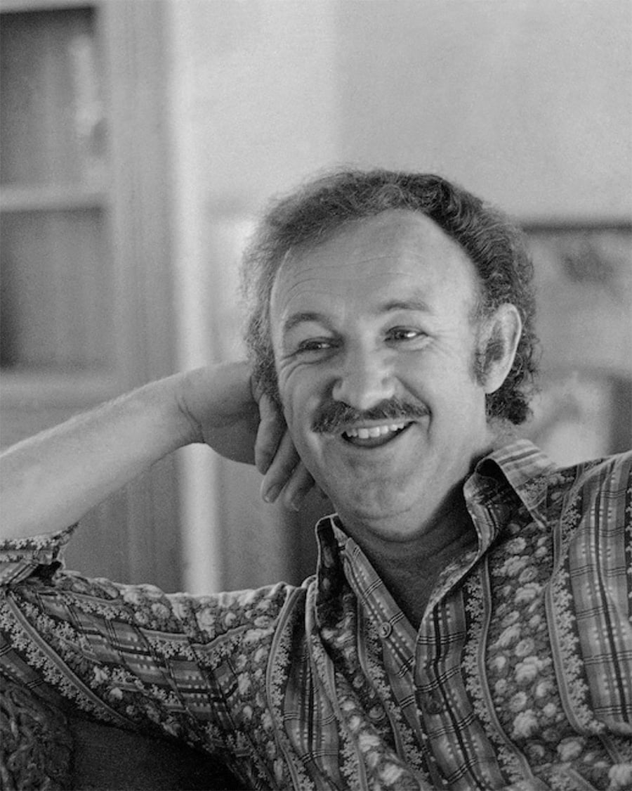 A man with curly hair and a mustache smiles while resting his head on his hand. He wears a patterned shirt and sits indoors, with shelves and a blurred background behind him. The photo is in black and white.
