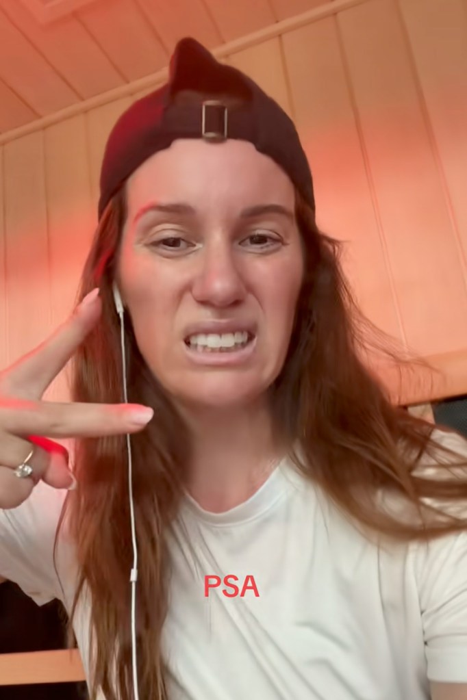 Comedian Hannah Berner making a sour face and wearing a black backward baseball cap, white t-shirt, and white wired earbuds.
