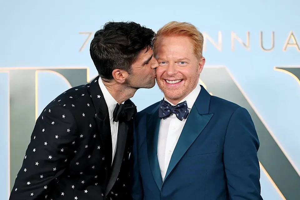 Two men smiling on a red carpet; one in a star-patterned suit kisses the cheek of the other in a teal suit with a bow tie