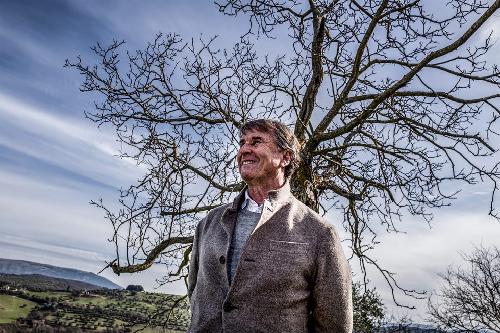 Smiling man standing outdoors next to a bare tree with a landscape of rolling hills in the background. Smiling man standing outdoors next to a bare tree with a landscape of rolling hills in the background.