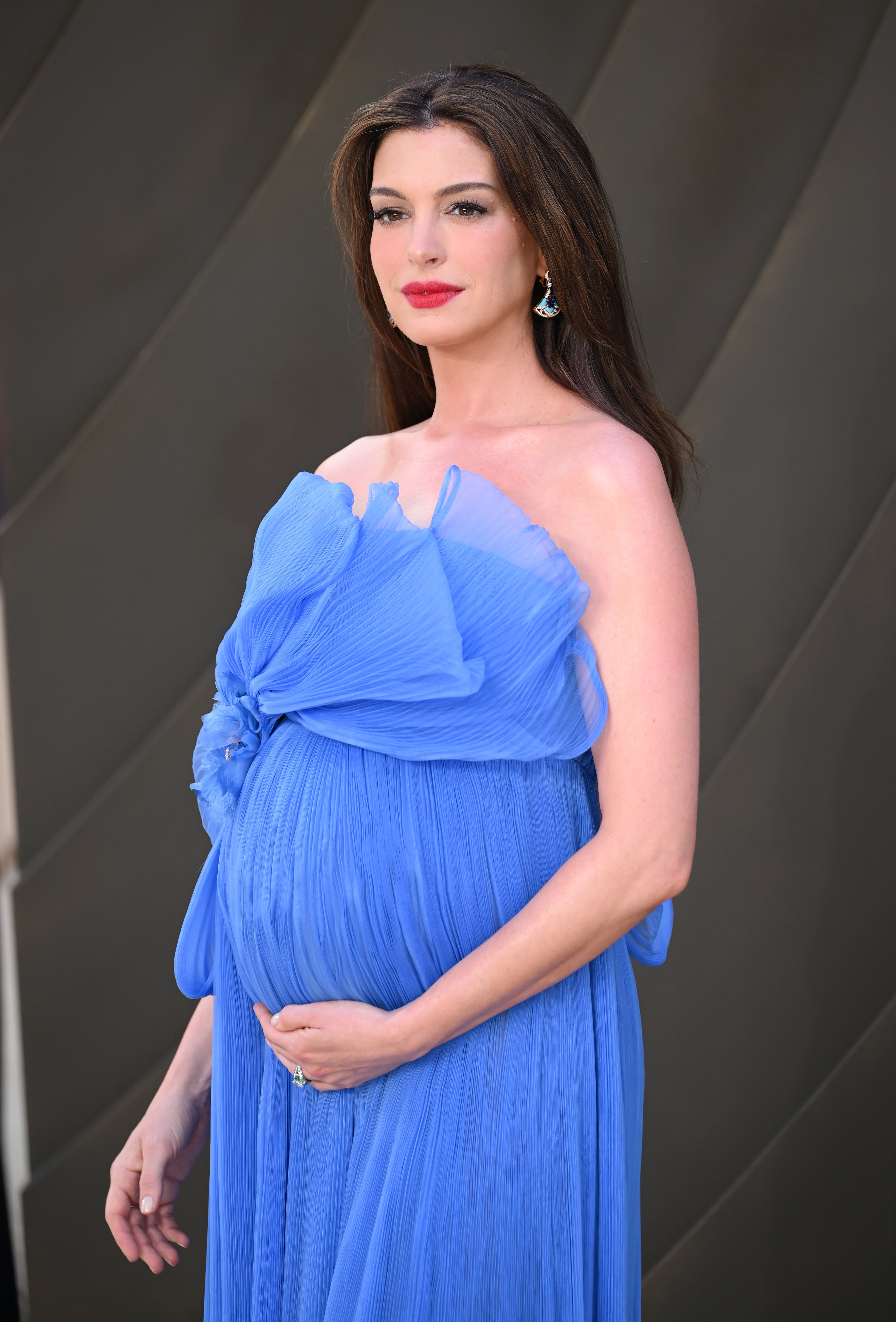 A person in a strapless, flowing gown with a large bow detail, cradling their baby bump, and wearing statement earrings on a formal event backdrop